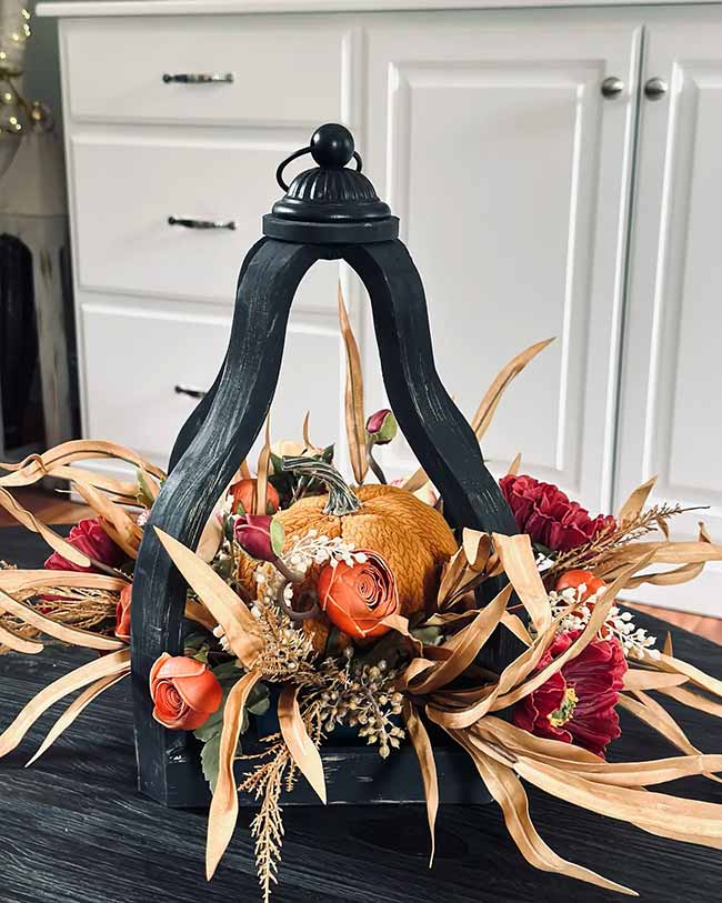 This dark, carved wood lantern is used as a centerpiece, filled with a small bumpy orange pumpkin and deep red and orange flowers. The dry golden leaves sticking out everywhere make it look super lush and ready for autumn.