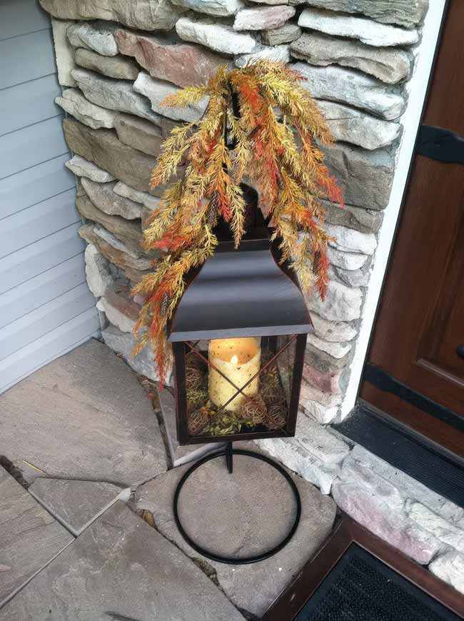 This black metal lantern is standing on a dark metal base right next to a stone wall. The candle inside is surrounded by small mossy balls, and the bundle of golden wheat tied to the top gives it a great, simple autumn look for the front door.