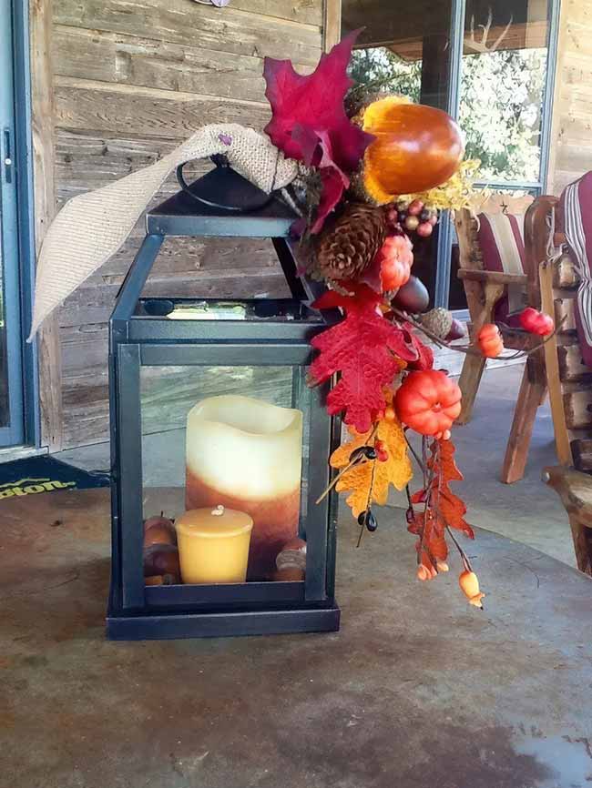 Wow, this large, black outdoor lantern is fully dressed up for the season! It has a giant two-tone candle inside, and the top is covered with a big burlap bow and a beautiful spray of red leaves, acorns, and small orange pumpkins hanging over the side.