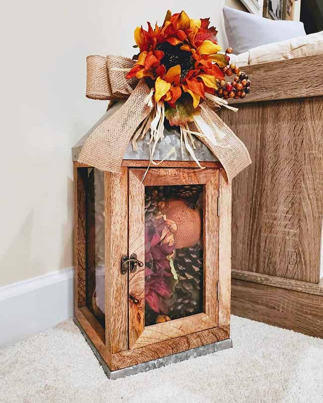 This tall wooden lantern has a cool metal base and top, and the inside is stuffed with pinecones, a small orange pumpkin, and dark red dried flowers. Tying a burlap bow with a bright sunflower on top is a classic rustic fall lantern design.