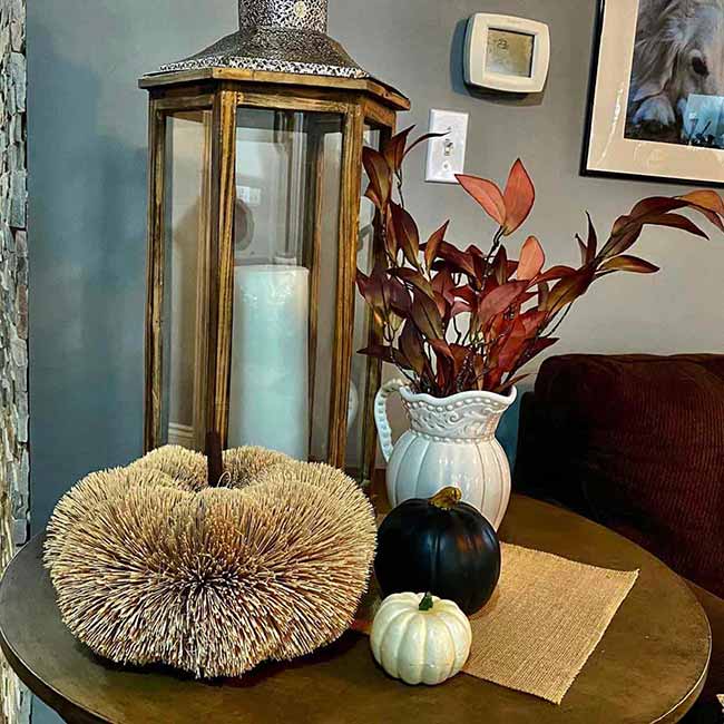 This tall wooden lantern with the white candle stands next to a vase of dark red leaves. The furry, round pumpkin on the table is an unexpected texture that adds a fun, cozy feeling to the whole arrangement.