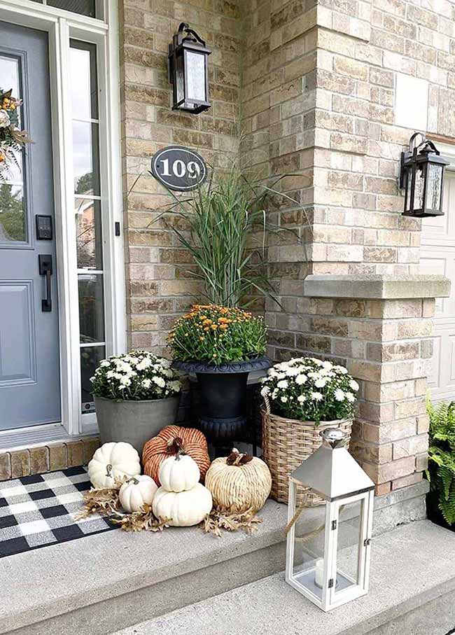 This bright white lantern with a glass top sits on a black and white checked rug next to several orange and white pumpkins. The flowers and tall grass make this a balanced, cozy front porch decor setup.