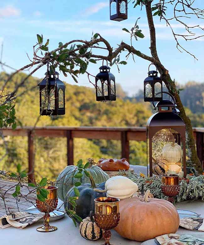 This outdoor dining table is decorated with beautiful gourds and pumpkins, and small black lanterns hang from the tree branches overhead. The large lantern on the table holds white pumpkins and vines, adding to the lovely natural fall decor theme.