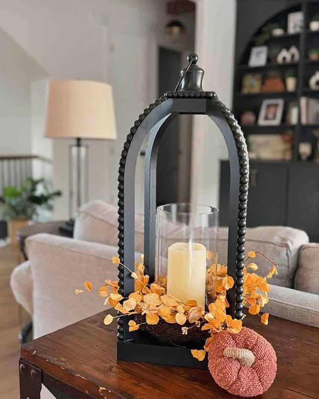 This black lantern with a curved, beaded top holds a single white candle surrounded by small orange berry sprigs. The little knitted pumpkin next to it gives the whole piece a warm, homemade fall centerpiece feel.