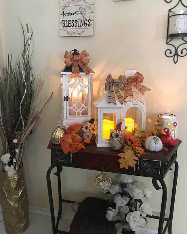 These two white painted lanterns on the console table glow with yellow candles and are topped with checkered bows. Arranging them with small white pumpkins and golden leaves makes this a very charming harvest blessing display.