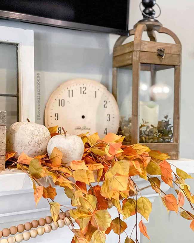 This rustic wood lantern on the mantel is surrounded by beautiful golden leaves and two small white pumpkins. The faded round clock face behind it gives the whole mantel decor arrangement a lovely vintage look.