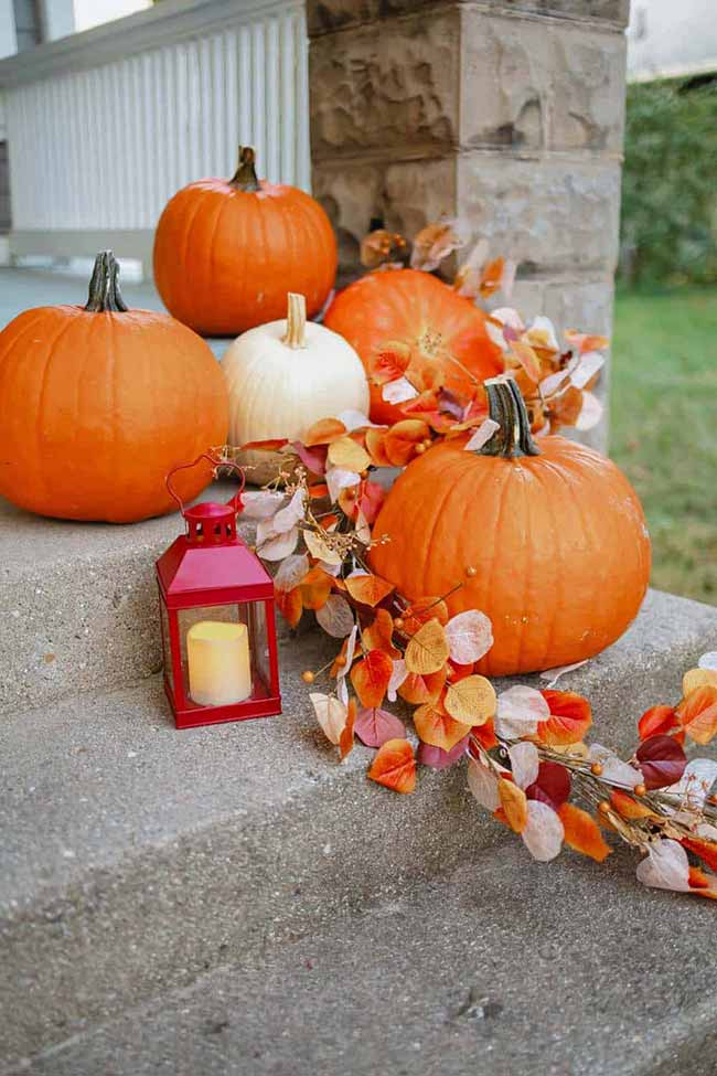 A tiny, bright red lantern with a single candle sits on the stone steps amongst large orange and white pumpkins. The colorful garland of autumn leaves draped over the stairs connects the whole simple welcome display.