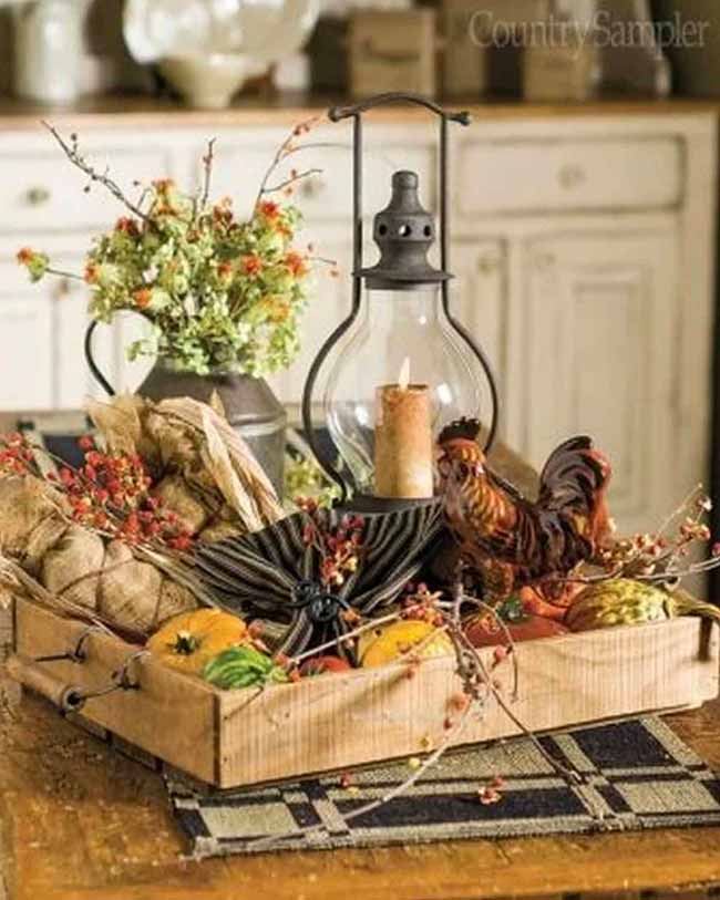 I love this charming centerpiece: a wooden tray holding a clear glass bubble lantern with a candle, surrounded by dried corn husks, pumpkins, and a little rooster figurine. The whole arrangement has a wonderful country sampler fall style.