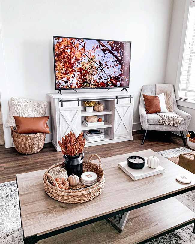 The white TV stand with the barn doors is a great farmhouse piece. A small tray holds mini pumpkins and dried flowers for a charming coffee table display. The comfy gray chair and brown pillows make this a cozy corner for watching TV.