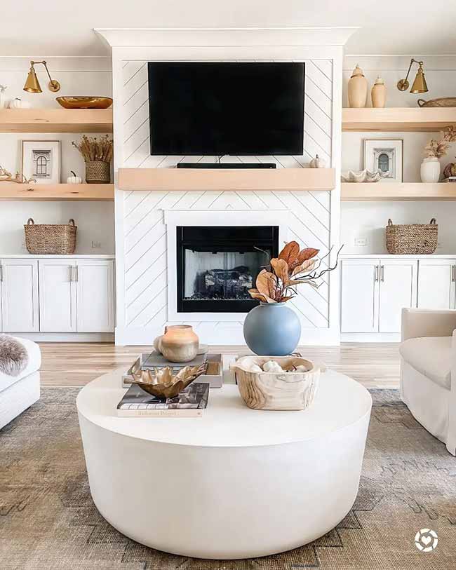 This living room is light and modern with a focus on clean lines. The open wood shelves next to the fireplace are decorated with simple fall pieces like small white pumpkins and dried wheat. The big round coffee table is super chic.