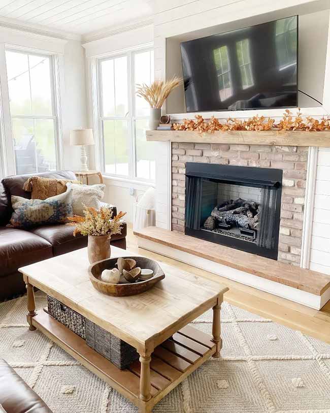 The fireplace has a beautiful light brick and a chunky wood mantel. A simple garland of orange leaves and a vase of dried wheat bring the season in. The big windows make the room feel bright and airy, even on a cloudy day.