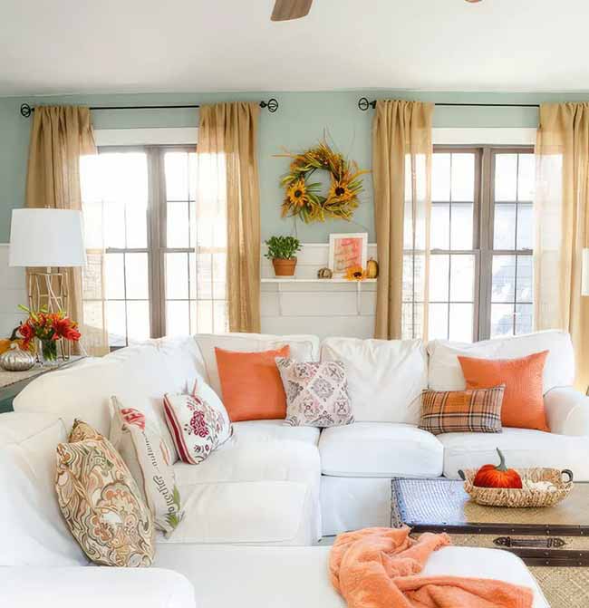That bright orange throw and pillows really pop against the white couch. The sunny color makes this living room feel happy and ready for fall. The big sunflower wreath above the shelf is a perfect touch.