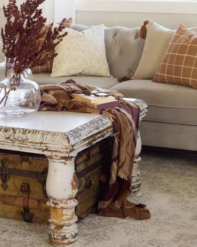 The rustic coffee table made from an old trunk is a clever idea for this farmhouse look. The rich brown blanket and dried red flowers match the warm colors of the tufted couch pillows. Everything here looks cozy and well-loved.