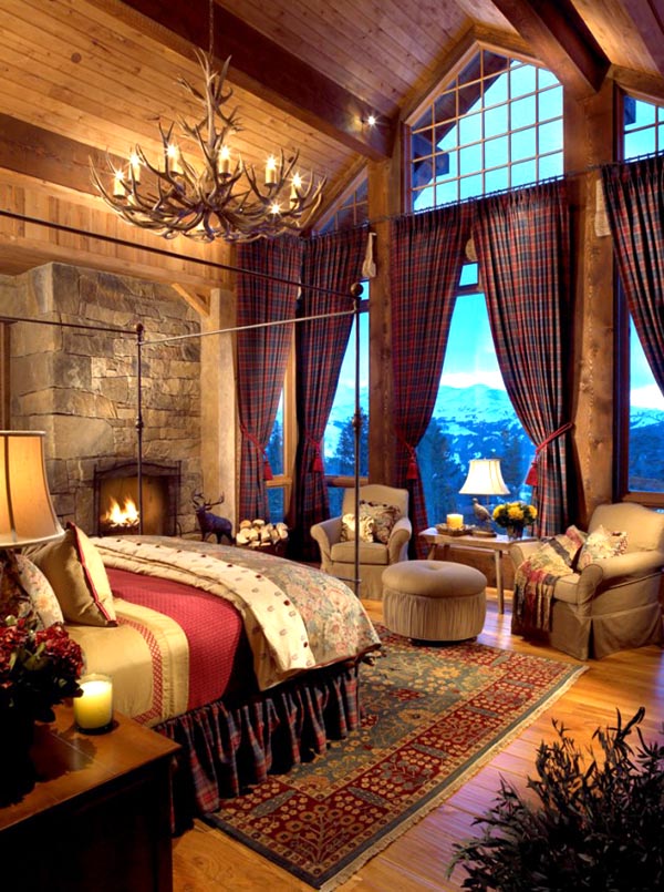 This grand master bedroom features a cozy, dedicated sitting area by the window, complete with two armchairs and an ottoman, perfect for enjoying the stone fireplace. The huge antler chandelier and rustic wood ceiling emphasize the warm, luxurious cabin atmosphere.