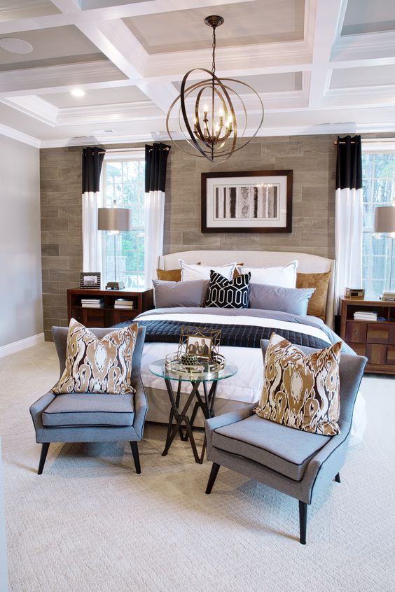 A stylish, central sitting area is created by two handsome gray armchairs and a small round table, positioned directly across from the bed. This is a perfect arrangement for a conversation, set against the striking stone accent wall.