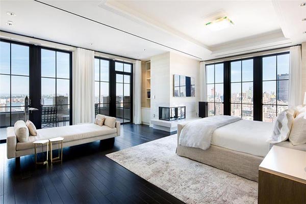 An impressive, sprawling master bedroom with an urban view features a long, modern white chaise lounge set by the floor-to-ceiling windows. This expansive sitting area is ideal for taking in the cityscape, complemented by the clean lines of the dark wood floor.