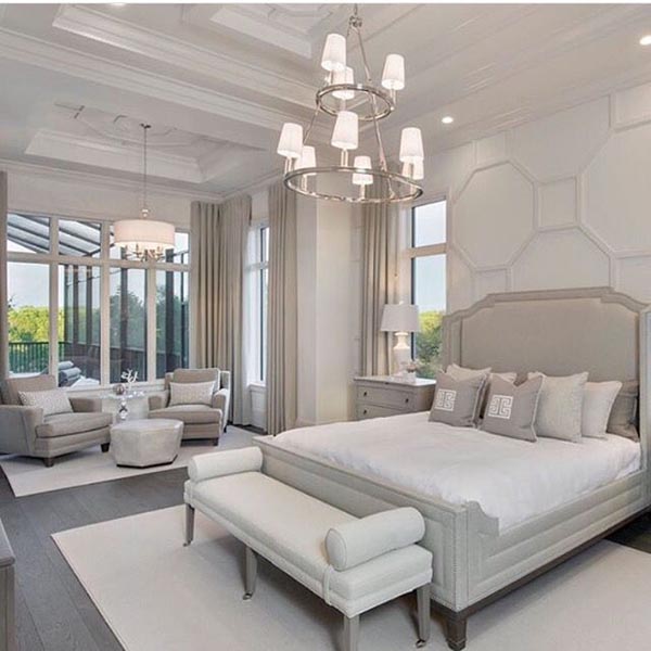 This luxurious master bedroom is designed with a spacious, separate sitting area by the large patio doors, featuring two soft gray armchairs and an ottoman. The elegant paneled accent wall and layered white rugs ensure the whole suite feels cohesive and serene.