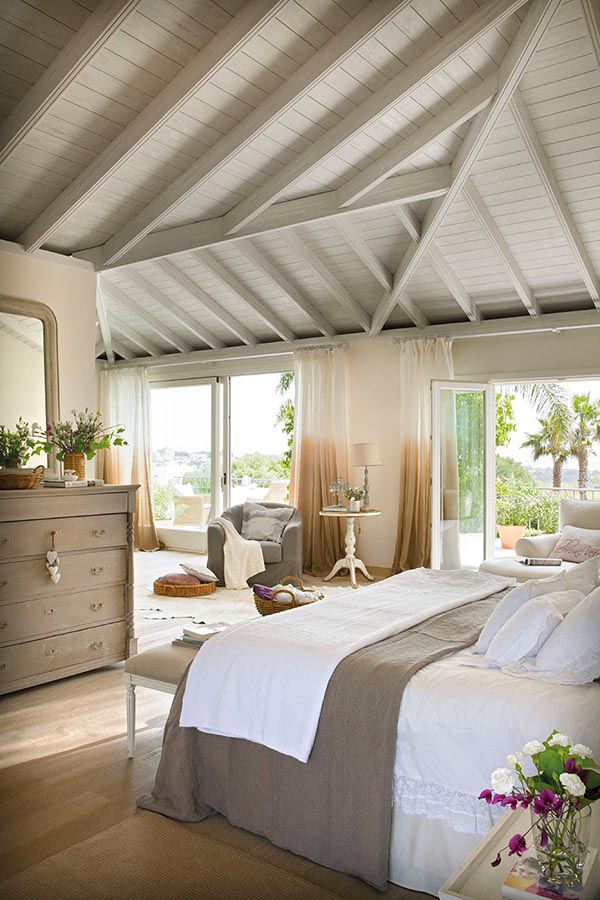 What a wonderfully bright and breezy master suite! The charming vaulted wooden ceiling creates a cottage feel, and a small, inviting sitting area is nestled by the doors with a gray armchair, perfect for enjoying the sunny view.