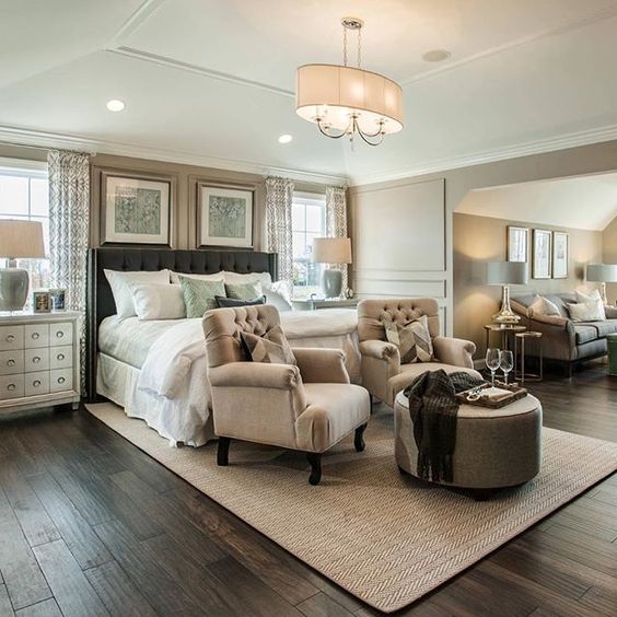 This expansive master bedroom uses a smart layout to define its sitting space. Two neutral tufted club chairs and a central ottoman on a chevron rug create an immediate conversation area, flowing seamlessly into the larger lounge visible in the background.