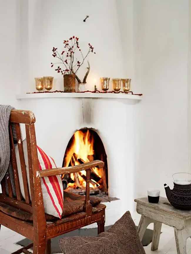 Fireplace Comfort. A small, cozy fire burns in this bright white fireplace. The shelf above it has just a few gold glasses and a small branch with red berries for a spot of color. This looks like the perfect spot for a relaxing afternoon read.