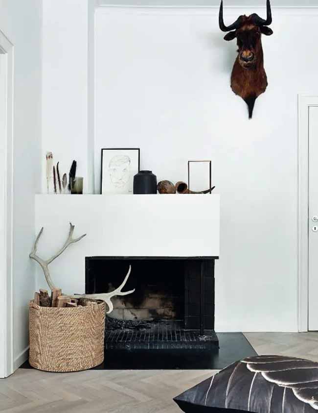 Taxidermy and Textures. The dark animal head on the white wall adds a cool, rustic feel to the room. The mantelpiece is kept clean with just a few natural items and simple art. The big basket of logs next to the black fireplace looks really cozy.