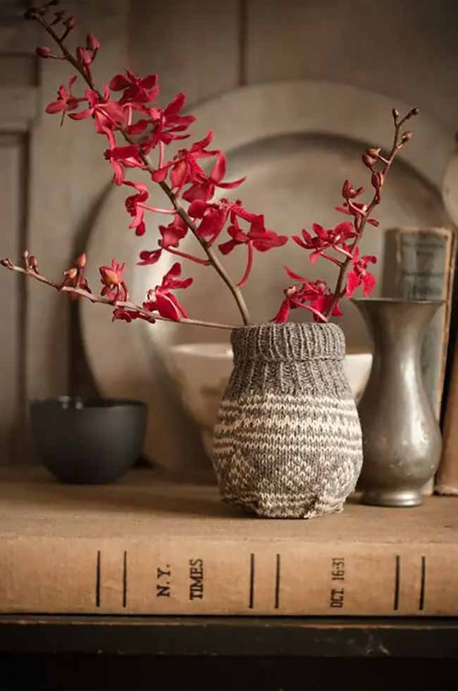 Knitted Flower Holder. This small vase has a cozy knitted cover with a gray and white pattern. The bright red flowers really stand out against the soft textures and old books underneath.