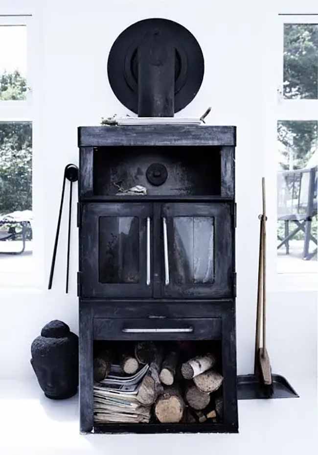 Black Stove Focus. This tall, black wood stove is the clear center of the room and holds a stack of logs and old papers underneath. The contrast between the dark stove and the bright white room is very striking and modern.