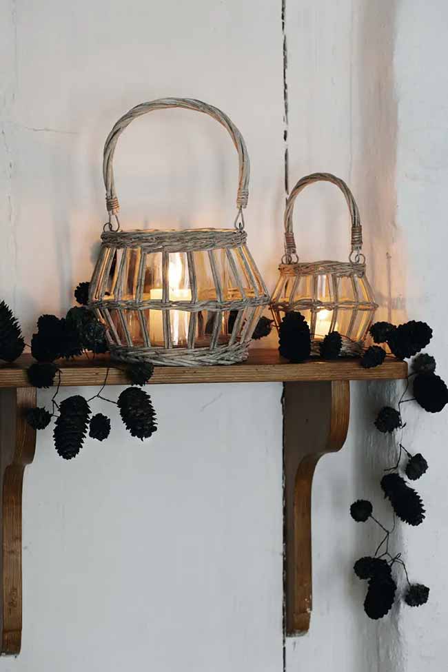 Wicker Lanterns. These small, charming lanterns are made of wicker and glass and sit nicely on a rustic wooden shelf. The dark black garland of pinecones hanging down adds a nice rustic contrast to the light wood.