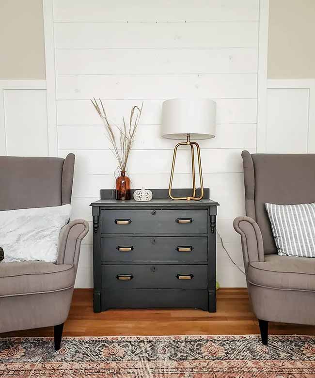 Symmetrical Reading Corner. Two gray armchairs flank a dark wooden chest, creating a formal, balanced look. The elegant gold lamp and natural dried grass stems add texture to the simple white wall.