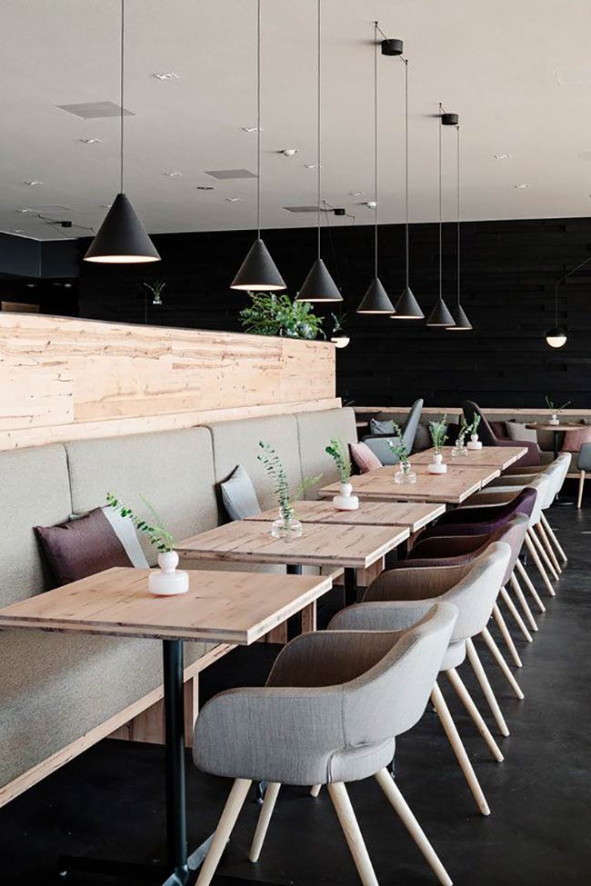 A long row of matching black cone lights hangs over the tables to create a clean line. The light wood furniture looks very simple and organized against the dark black wall.