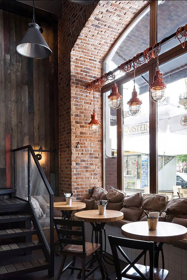 Red cage lights hanging by the big window add a nice pop of color to the raw brick wall. The high arched ceiling and metal stairs give this coffee shop interior a strong industrial look that feels very hip and urban.