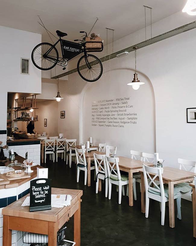 The high white ceiling gives the room an airy, gallery-like feeling. A bicycle hanging overhead acts as a central art installation. White tables and chairs below are classic and bright.