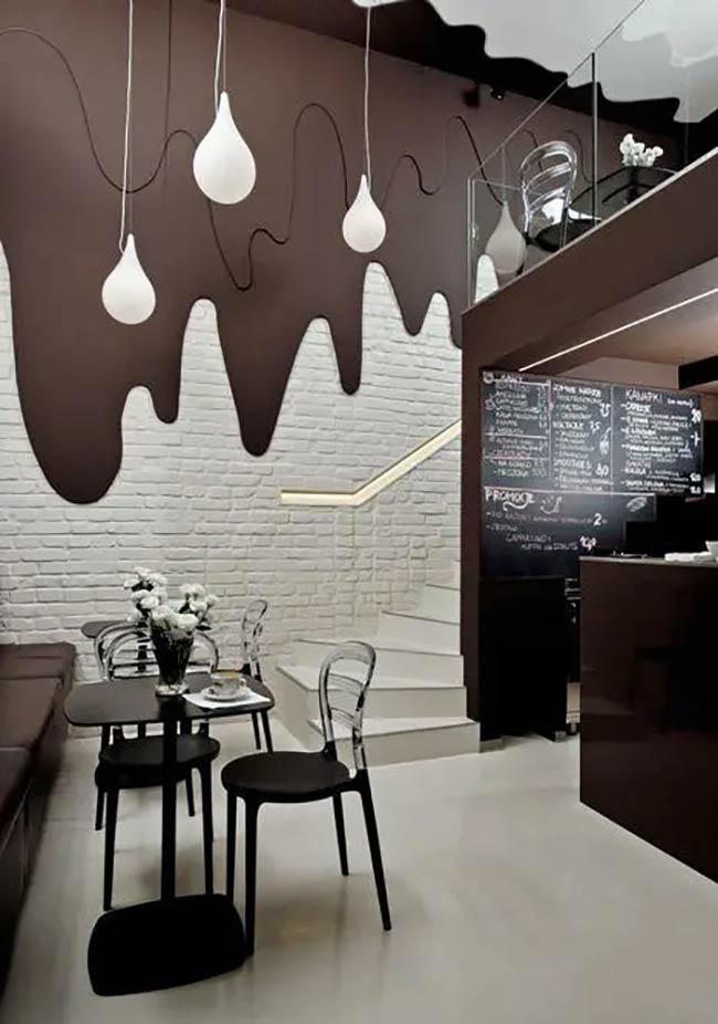 The dramatic wall features a chocolate-drip texture over white subway tiles. White, teardrop-shaped pendant lights hang from the ceiling, mimicking falling drops. This dark, playful theme continues with transparent black chairs and a chalkboard menu.