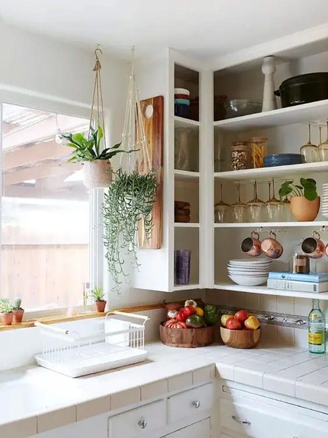 Open white shelves wrap right around the corner to use every bit of space. Hanging green plants and shiny copper mugs make the room feel bright and happy. It is a great no door kitchen cabinet solution for a sunny kitchen window.