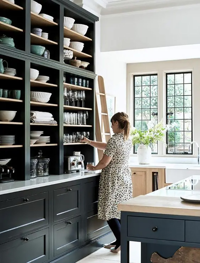 This huge dark cabinet covers the whole wall like a library bookcase. A wooden ladder helps you reach the high bowls and jars on the top shelves. The dark color makes the white dishes pop in this dramatic no door kitchen cabinet display. It creates a stunning focal point for the room.