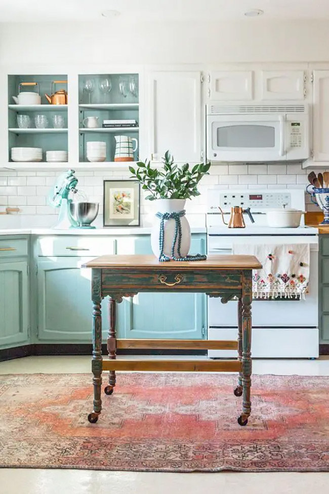 White open shelves on top look great with the teal cabinets on the bottom. An old wooden table in the middle adds a rustic touch to the colorful room. This no door kitchen cabinet layout mixes old and new styles beautifully.
