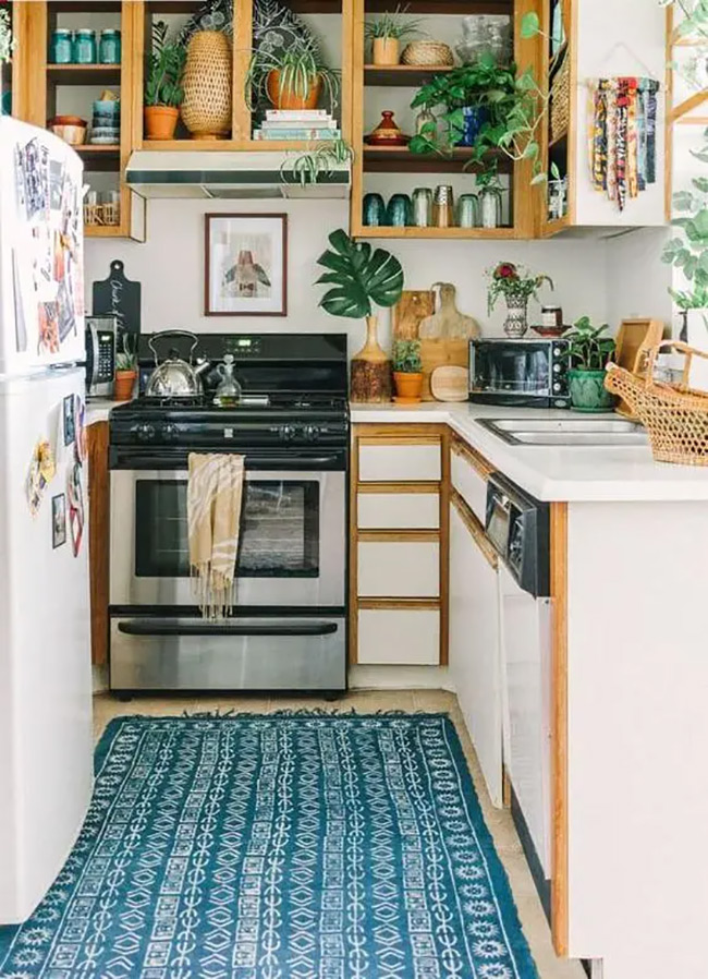 Natural wood cabinets with no doors add warmth to this kitchen. They are filled with plants, colorful jars, and cookbooks, giving the room a lively and lived-in feel. The blue patterned rug adds a nice splash of color on the floor.
