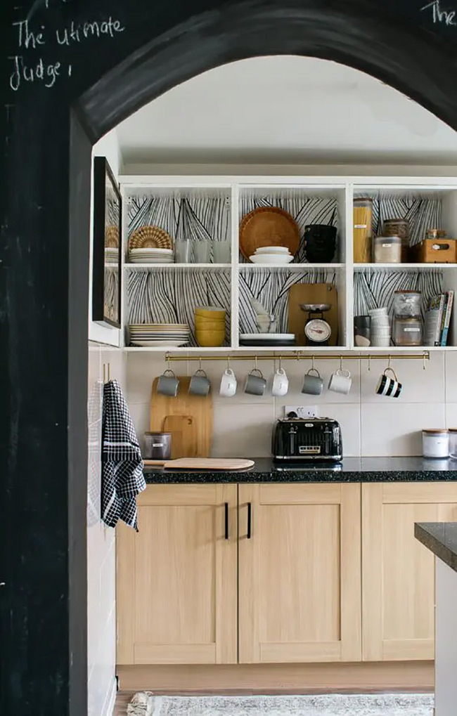 Black and white patterned wallpaper lines the back of these open white cabinets. It makes a striking background for the simple white dishes and wooden bowls. This artistic touch turns storage into a decor feature.