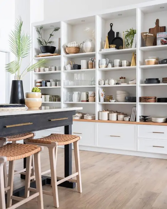 A massive wall of white shelves provides incredible storage space for dishes, jars, and decorative items. The bottom drawers offer hidden storage for less pretty things. It looks like a professional chef's pantry right in the kitchen.