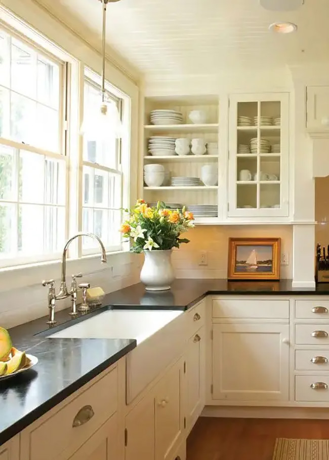 Simple white shelves are built into the wall next to the bright window. They hold stacks of white dishes that blend in perfectly with the white beadboard. It is a very clean and classic no door kitchen cabinet look.