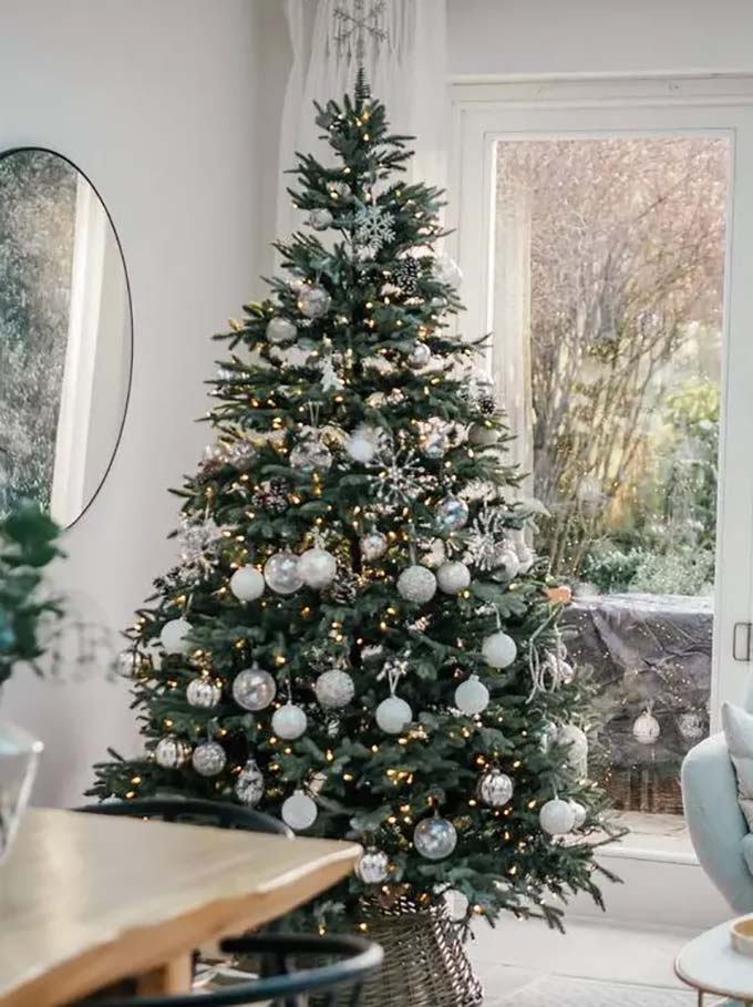 A big green tree looks very traditional standing by the glass door. It's decorated simply with white balls, sparkly snowflakes, and silver ornaments.
