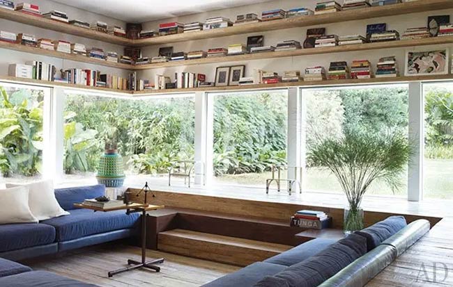This sunken living room is surrounded by windows that look out into a green garden. Wooden steps lead down to the blue sofa. Books are stored on high shelves all around the room.