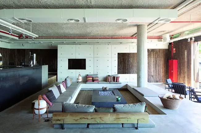 That's a very industrial-looking room with all the concrete and red pipes on the ceiling. A simple gray sofa sits in the sunken conversation pit, which has a fire pit in the middle.