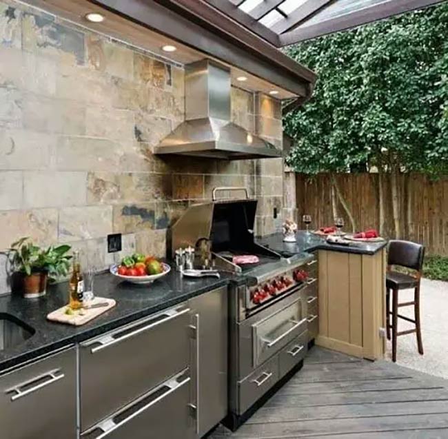 This setup looks like a professional kitchen with that massive stainless steel grill and hood. The natural stone tiles on the wall add a warm touch to the modern outdoor barbeque area. It sits nicely on the wood deck under a protective roof.