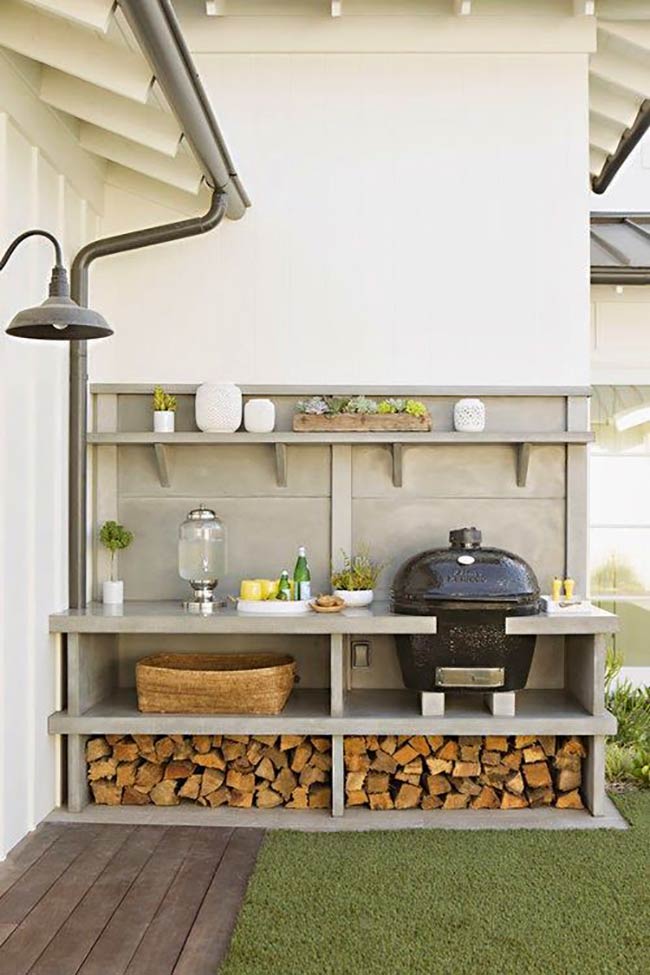 A  grill sits next to the regular gas barbecue for serious cooking. The concrete countertop provides plenty of space for prepping food, while wood storage underneath keeps logs dry. It’s a lovely farmhouse-style setup on a grassy patio.
