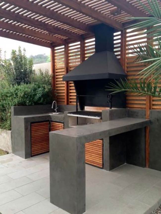 A long concrete bar extends from the grill station, offering a great place to stand and chat. The dark wood pergola with slats lets sunlight filter through while defining the cooking zone. It looks very solid and modern.