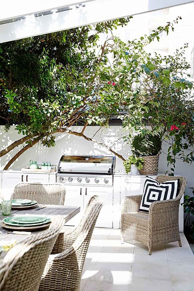 White brick cabinets look bright and fresh under the shade of large trees. The built-in grill is ready for action next to comfortable wicker dining chairs. This outdoor barbeque area feels like a cozy extension of the home.