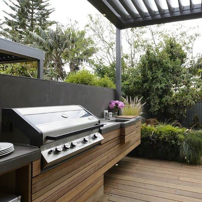 This sleek kitchen uses warm wood slats and cool grey concrete to create a stylish look. The sink is built right into the counter next to the large grill. It sits under a modern roof that lets in just enough light for cooking.