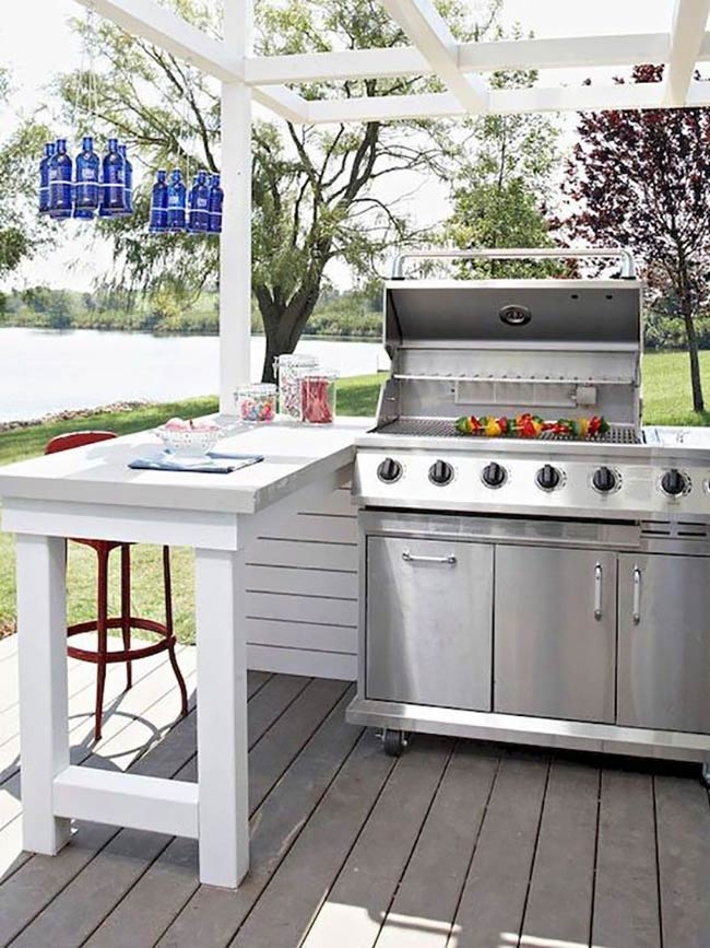 A white wooden island creates a simple prep space right next to the big stainless steel grill. Blue bottles hanging as a light fixture add a fun pop of color. The view of the water makes this a relaxing place to cook.