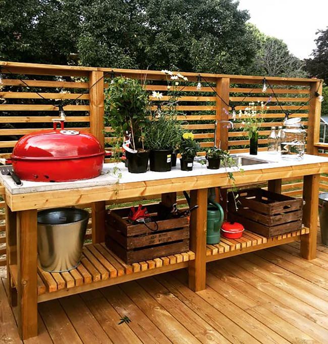 A simple wooden workbench serves as a great station for the bright red kettle grill. String lights hang from the privacy screen to set a cozy mood. Crates on the bottom shelf hold all the necessary tools for a cookout.